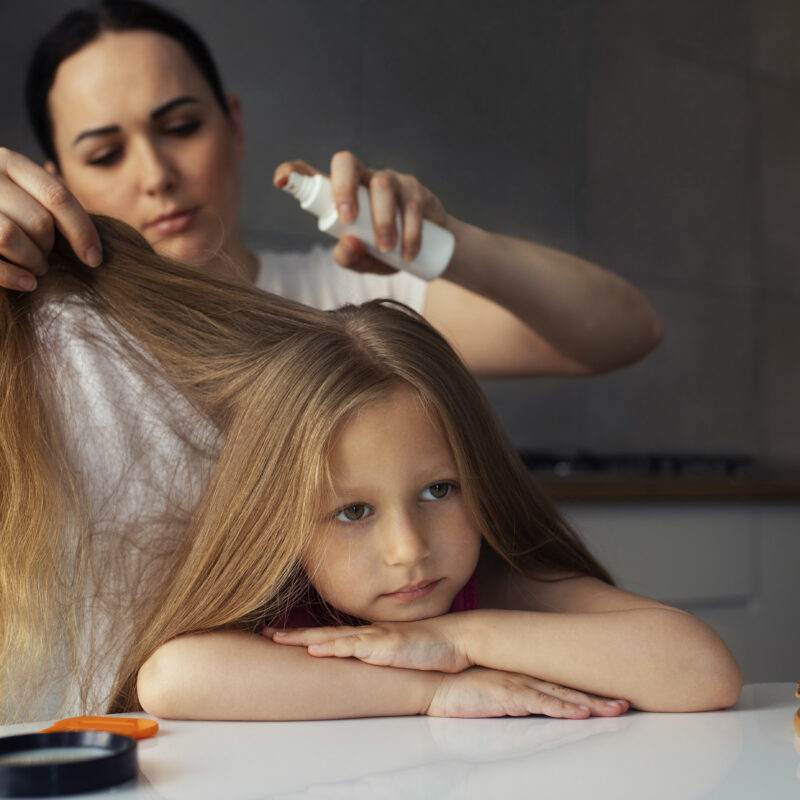 A woman sprays a hair treatment onto her daughter's long brown hair. The girl is sitting at a table.